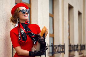 Beautiful woman in red beret and dress walks down the street of city, black gloves, dark glasses. Attractive beauty girl French style with scarf around her neck. Holding package baguette of bread